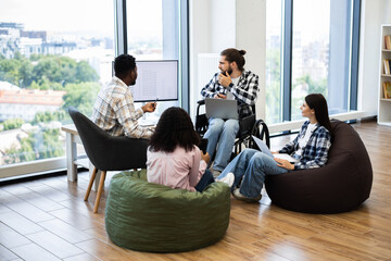 Diverse group of young professionals collaborates in casual office with inclusive workspace setting. Individuals focus on discussion and development of plans, fostering teamwork and creativity.