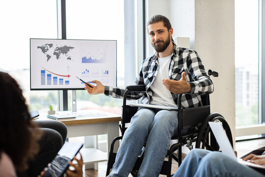 Young male presenter in wheelchair discussing economic report with professionals, highlighting trends using chart on monitor. Business presentation emphasizes diversity inclusion in modern workplace. - Powered by Adobe