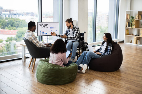 Diverse professionals discussing financial report in office. Includes young wheelchair user leading conversation and mixed gender team interacting around monitor with graphs.
