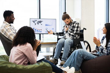 Diverse team including man in wheelchair discuss economic challenges using charts and graphs, showcasing effective collaboration, teamwork, inclusivity in office, inspiring professional environment.