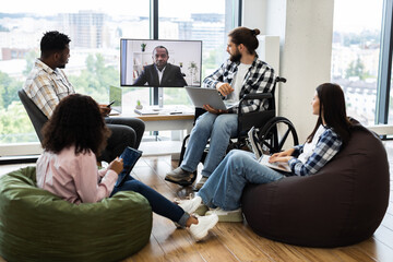 Group of young professionals discussing with team leader through a video call in open workspace featuring natural light and inclusive seating including bean bags and wheelchair.