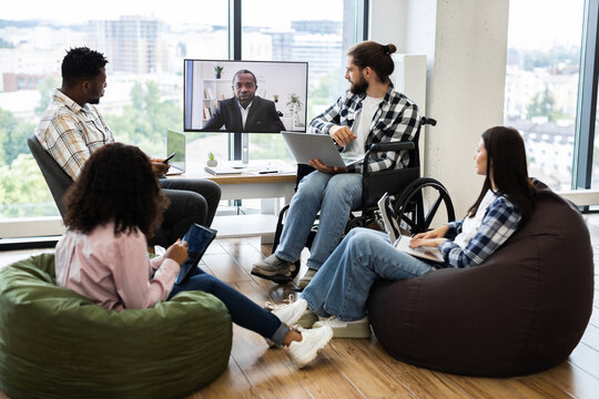 Group of young professionals discussing with team leader through a video call in open workspace featuring natural light and inclusive seating including bean bags and wheelchair.