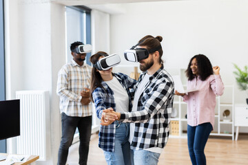 Diverse group of young professionals enjoying activities with virtual reality glasses inside office setting. Two dancing while others laugh, demonstrating happiness and creativity in technology.