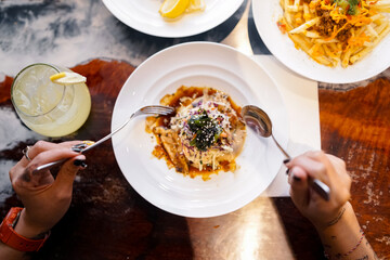 A person holds a fork and spoon eating okonomiyaki from a white plate, with a cold lemon drink on...