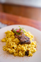 A plate of yellow fried rice topped with grilled beef slices, pickled shallots, and green onion garnish, served on a white dish over a modern patterned table.