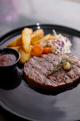 A grilled steak garnished with rosemary and garlic, served with golden potato wedges, fresh coleslaw, mushrooms, tomato slices, and a side of savory sauce on a black plate.