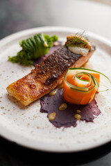 A gourmet dish with crispy-skinned salmon fillet, served with purple yam puree, carrot and cucumber ribbons, green vegetable, and a dollop of cream topped with fresh herbs.