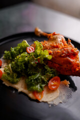 A grilled quail coated in spicy herbs, served with leafy lettuce, sliced chili, and a dollop of creamy sauce on a black plate for a gourmet presentation.