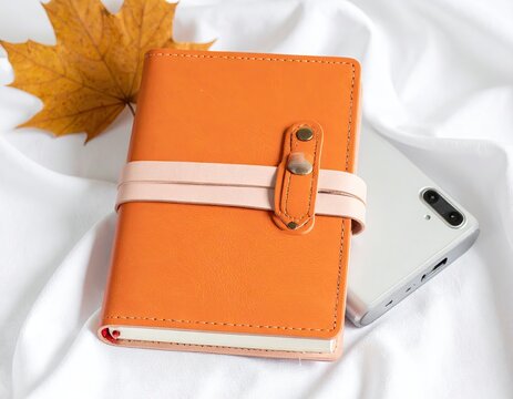 Orange notebook and smartphone with autumn leaf on white fabric flat lay.