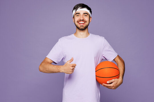 Young strong fitness trainer instructor sporty man sportsman in t-shirt training in home gym hold ball play basketball show thumb up isolated on plain purple background Workout sport fit abs concept