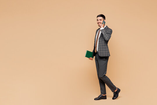 Full body young employee business man corporate lawyer wear classic formal grey suit shirt tie glasses work in office hold book talk speak on mobile cell phone walk isolated on plain beige background.