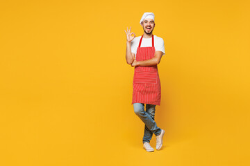 Full body young smiling happy housewife housekeeper chef cook baker man wear red apron toque hat show ok okay gesture isolated on plain yellow orange background studio portrait. Cooking food concept.