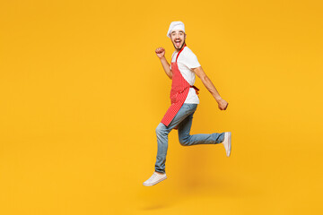 Full body young fun housewife housekeeper chef cook baker man wear red apron toque hat run jump high do winner gesture isolated on plain yellow orange background studio portrait. Cooking food concept.