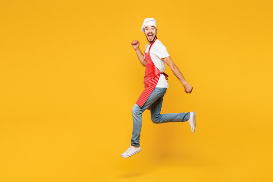 Full body young fun housewife housekeeper chef cook baker man wear red apron toque hat run jump high do winner gesture isolated on plain yellow orange background studio portrait. Cooking food concept.