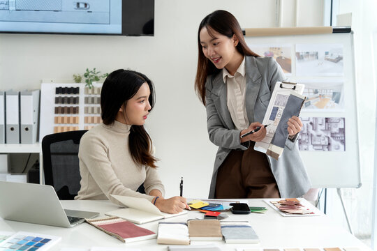 Asian adult women designers collaborating interior architectural plans fabric samples modern design studio office workspace material selection creative project.
