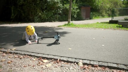 Toddler falling off scooter on park path wearing yellow helmet, playful accident during outdoor ride, moment of imbalance and spontaneity in carefree childhood adventure