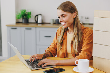 Young happy housewife IT woman wear orange shirt casual clothes hold use work on laptop pc computer sit at table near stack of cardboard boxes in kitchen at home alone. Lifestyle cooking food concept.