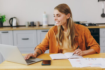 Young smart housewife IT woman wear orange shirt casual clothes hold use work on laptop pc computer write down in notebook sit at table in light kitchen at home alone. Lifestyle cooking food concept. © ViDi Studio