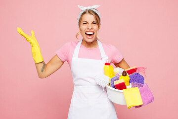 Young dissatisfied displeased woman wear white apron hold basin with detergent bottles while doing...