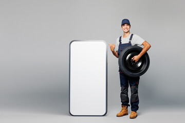 Full body young car mechanic man wear overall uniform workwear clothes work in garage point on big huge blank screen mobile cell phone isolated on plain grey background. Automotive repair job concept.