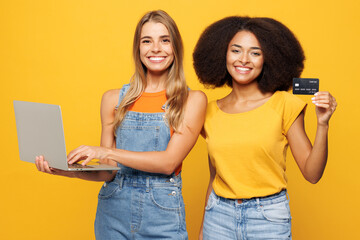 Two young friends buddies IT women wear casual clothes together using laptop pc computer hold credit bank card shopping online order delivery booking tour isolated on plain yellow orange background.