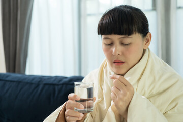 Sick female taking medicine drinking with water glass sitting under the blanket on sofa at home. Closeup woman holding pills capsule time to take medications. Healthcare concept.