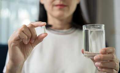 Sick female taking medicine drinking with water glass sitting on sofa at home. Closeup woman holding pills capsule time to take medications. Healthcare concept.