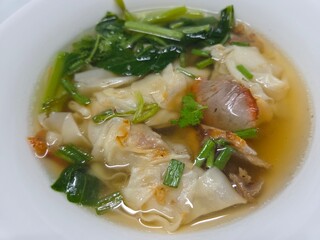 Close-up of a white bowl of wonton soup with folded wontons, sliced red roasted pork, green leafy vegetables, and chopped scallions in clear golden broth.