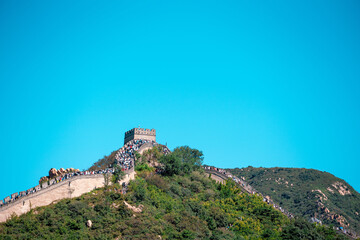 Obraz premium Ancient, weathered stone bricks of the Great Wall snake across a green, hilly landscape near Beijing, revealing tourists exploring the historical landmark under a clear sky.
