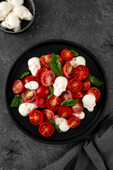 Halloween food. Caprese salad with mozzarella cheese skulls, basil and balsamic dressing on a black plate on a dark background. Funny food idea.