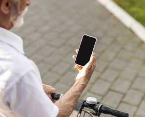 A joyful senior man interacts with his smartphone, standing adjacently to his bicycle, clad in casual attire indicative of leisure. Person using smartphone outdoors near bicycle on bright sunny day.