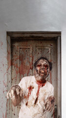 Scary male zombie with a wound and bloody standing in front of the old wooden door with splatters of blood drops on it. The scene is scary and creepy. Concept of Spooky Halloween