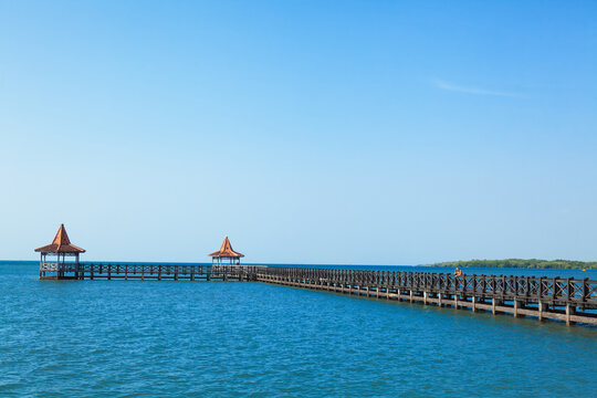 People walk by long wooden pier with traditional gazebos stretches over calm turquoise sea under clear blue sky. Perfect tropical scenery for travel, resort, summer vacation, coastal tourism themes.