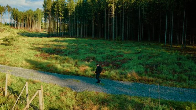 Backpacker walking on narrow gravel path by forest edge during sunny day. Explorer strolling near tree line under clear blue sky. Tourist moving through bright green landscape along woodland track