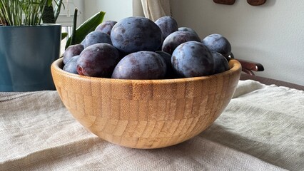 A wooden bowl filled with ripe plums sits on a light-colored fabric, with indoor plants and wooden coat hangers in the background.