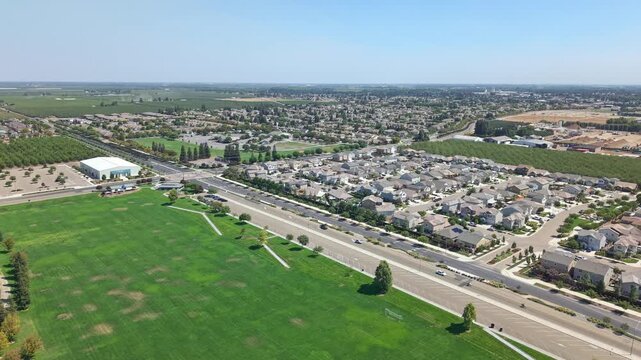 High drone shot rotating above grassy park and road dividing Ripon residential area, showing houses, traffic, farmland, and large recreational building.