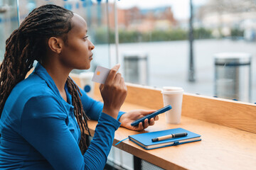A young woman is thoughtfully considering an online purchase, utilizing her credit card while sitting in a cafe