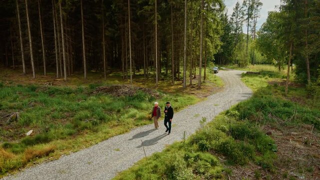 Woman walking next to grown son on forest trail in sunny weather. Gentleman strolling with elderly mother along gravel path under clear sky. Lady and man hiking through peaceful woodland road