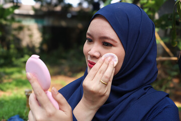 Young Muslim woman wearing hijab applying makeup with compact powder while looking in mirror...