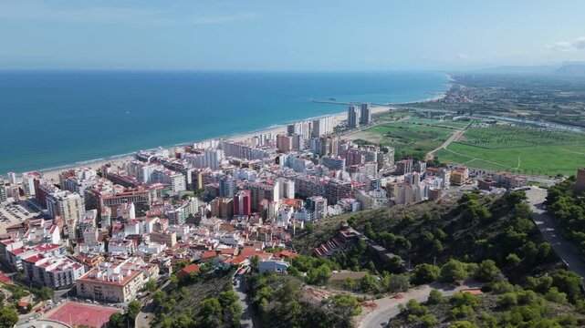 Aerial view of Cullera coastal cityscape with beachfront high-rises, Mediterranean Sea, green farmlands, and winding mountain roads in Valencia, Spain