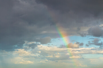Soft pastel rainbow shining through rain clouds with pale blue sky and gentle light, creating a delicate and atmospheric weather scene.
