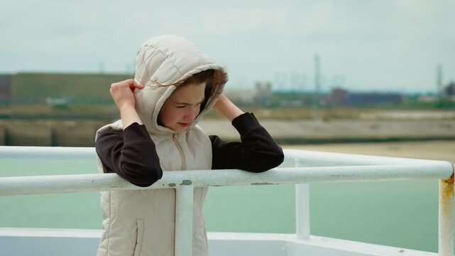 Girl standing on ferry deck during windy weather in Dunkirk port France. Child holding hood while feeling breeze near harbor railings. Young passenger enjoying boat ride on gusty day through coastal