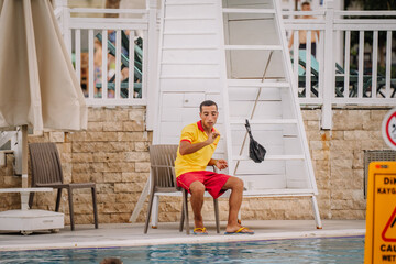 A lifeguard in a yellow and red uniform sits poolside on a chair near a lifeguard stand, watching the pool attentively with safety signs and swimmers nearby.