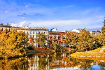 Beautiful autumn cityscape with colorful trees reflected in the water and historic buildings in the background. Calm lake with golden foliage and blue sky in a peaceful urban park. 