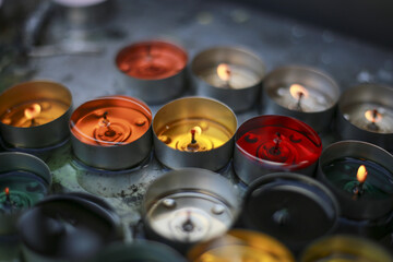 Colorful votive candles radiate soft glowing flames, casting warm light. Spiritual group, often...