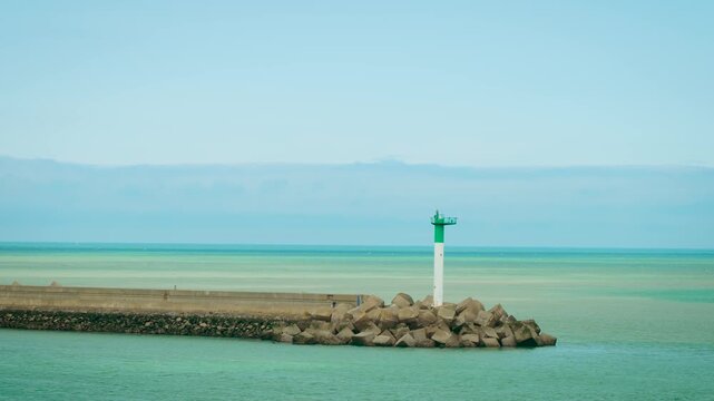 Green and white lighthouse standing on breakwater at Dunkirk port exit France. Beacon guiding vessels into calm turquoise sea under clear sky. Navigation light marking safe passage from harbor to open