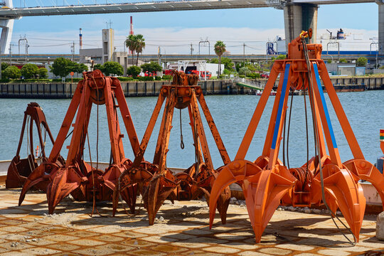 Dredging grab buckets used for harbor construction are lined up on the dockside at the port of Nagoya, Japan, with an industrial bridge and waterway in the background.