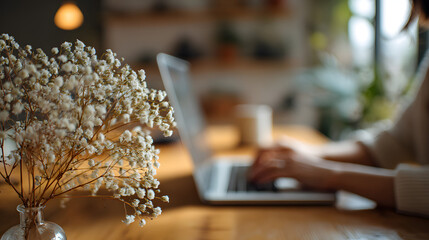Cozy Workspace: A serene scene capturing a person working on a laptop in a warm home setting with soft light, a vase of flowers adding a touch of nature, evoking a sense of focus and tranquility.