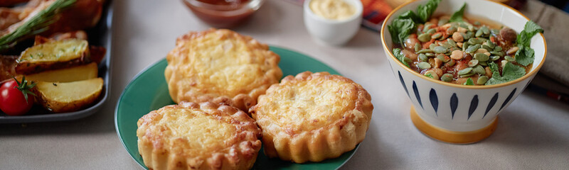 Website header shot of traditional Kwanzaa meal with three cheese tarts on plate next to bowl of soup garnished with pumpkin seeds and greens, side dishes visible in background
