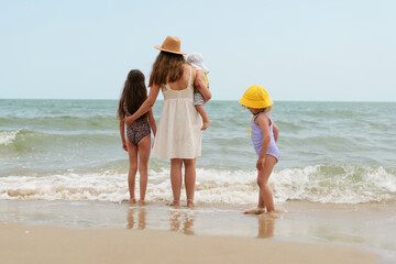 A mother stands with her children at the beach, watching the gentle waves roll in as they enjoy a sunny day by the ocean © sementsova321